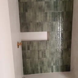 Ściana wykończona błyszczącymi, podłużnymi, zielonymi płytkami w łazience. Widoczna wnęka i złota bateria. Minimalistyczny odpływ liniowy w podłodze.