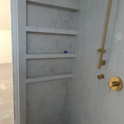 Wykończona łazienka z szarymi płytkami, wnęką z półkami i złotą armaturą. Widoczny fragment drzwi wejściowych. Minimalistyczny design.