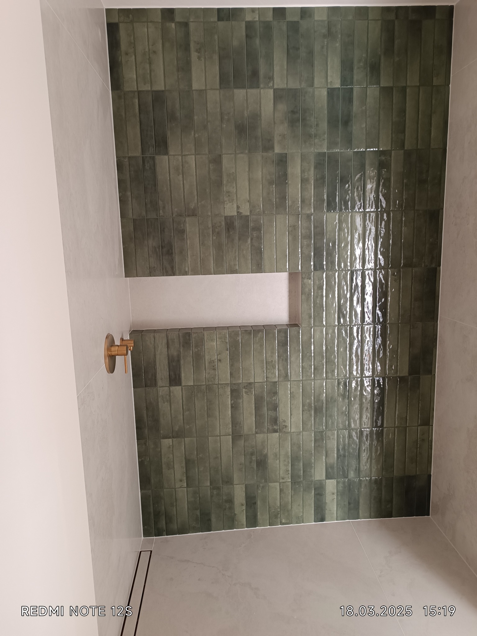 Ściana wykończona błyszczącymi, podłużnymi, zielonymi płytkami w łazience. Widoczna wnęka i złota bateria. Minimalistyczny odpływ liniowy w podłodze.