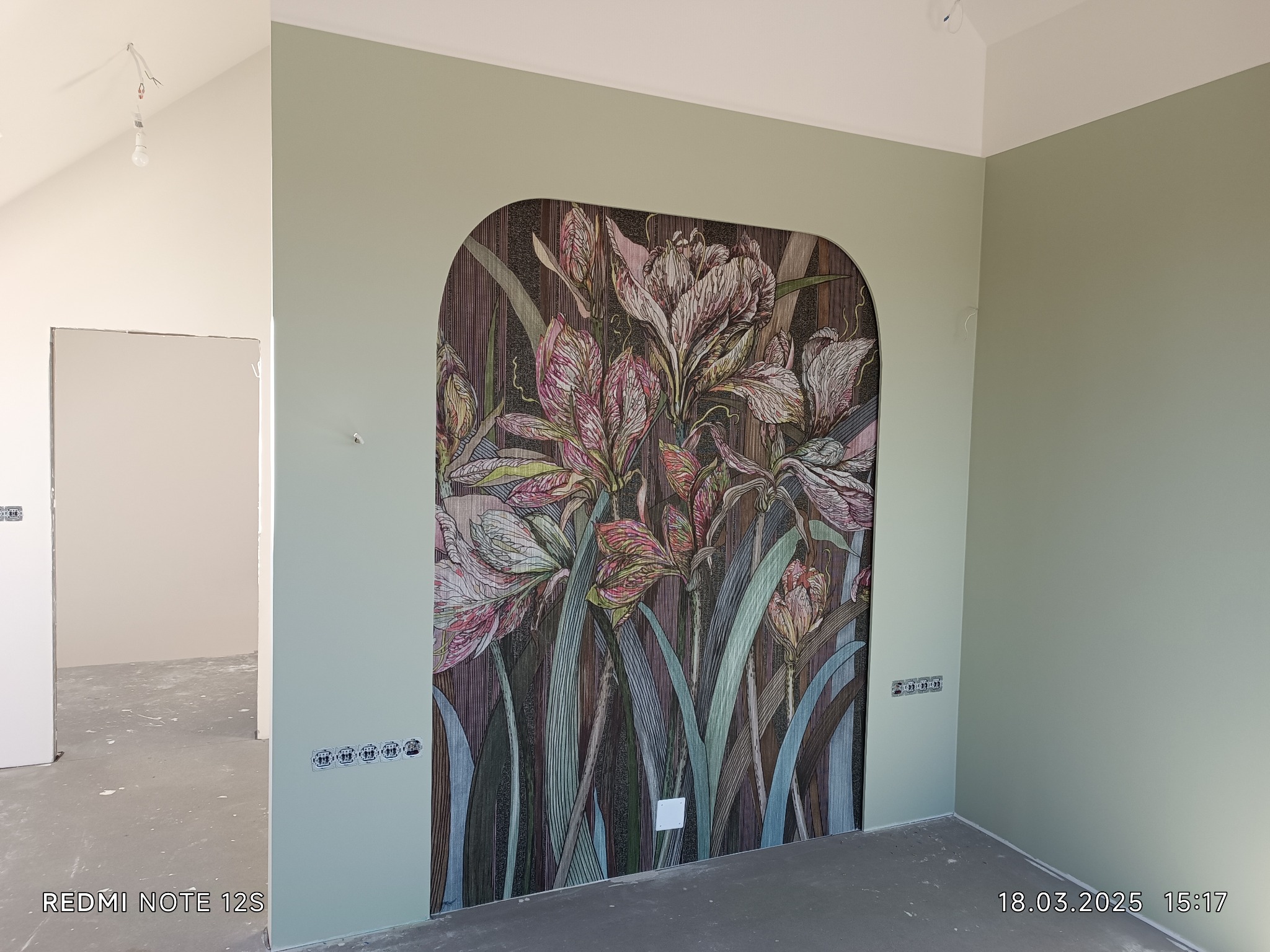 Dekoracyjna mozaika ścienna z motywem kwiatowym w łukowej wnęce, na tle świeżo pomalowanych ścian w Myślenicach. Widoczne gniazdka elektryczne i surowy stan podłogi.