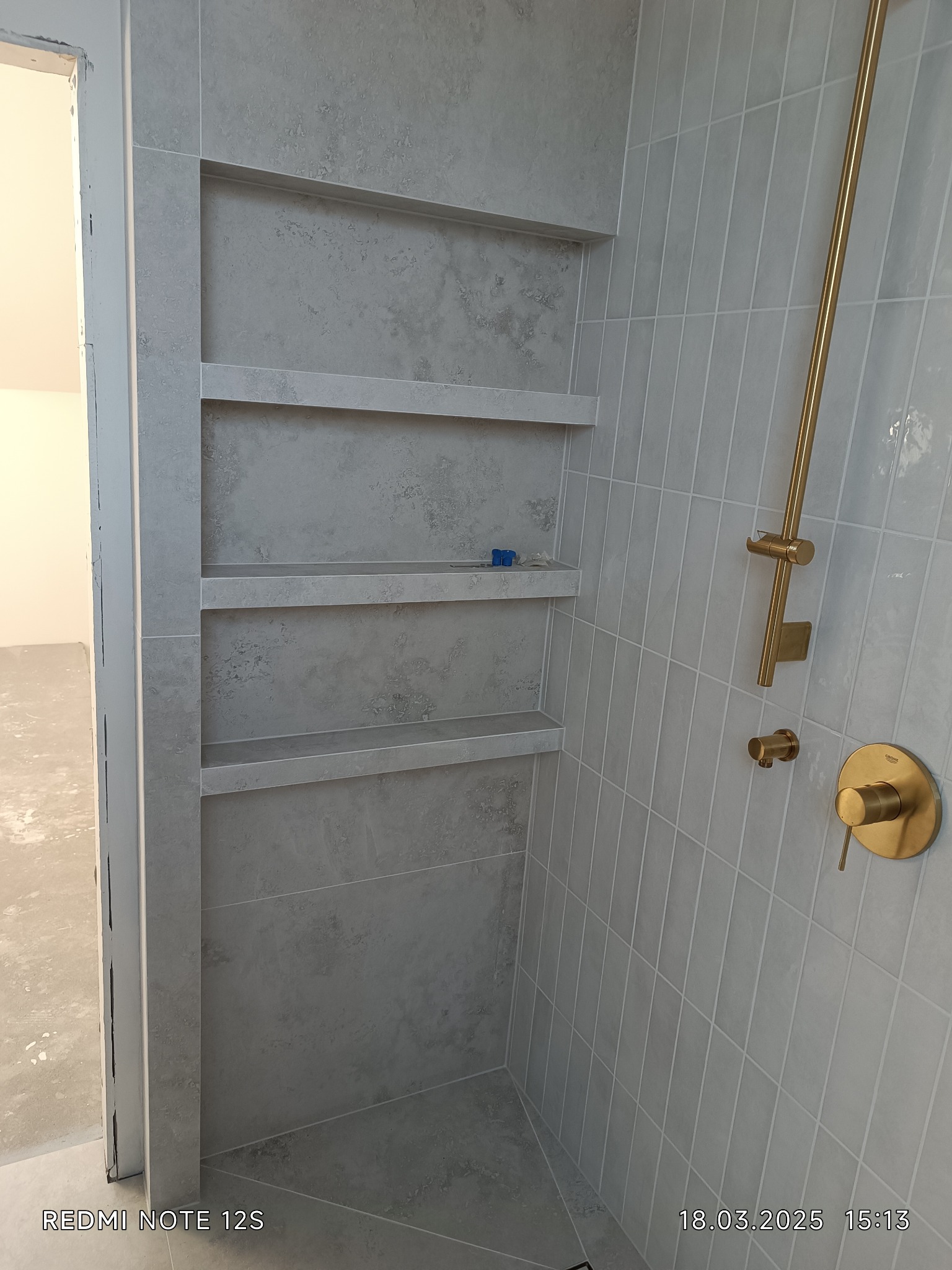 Wykończona łazienka z szarymi płytkami, wnęką z półkami i złotą armaturą. Widoczny fragment drzwi wejściowych. Minimalistyczny design.