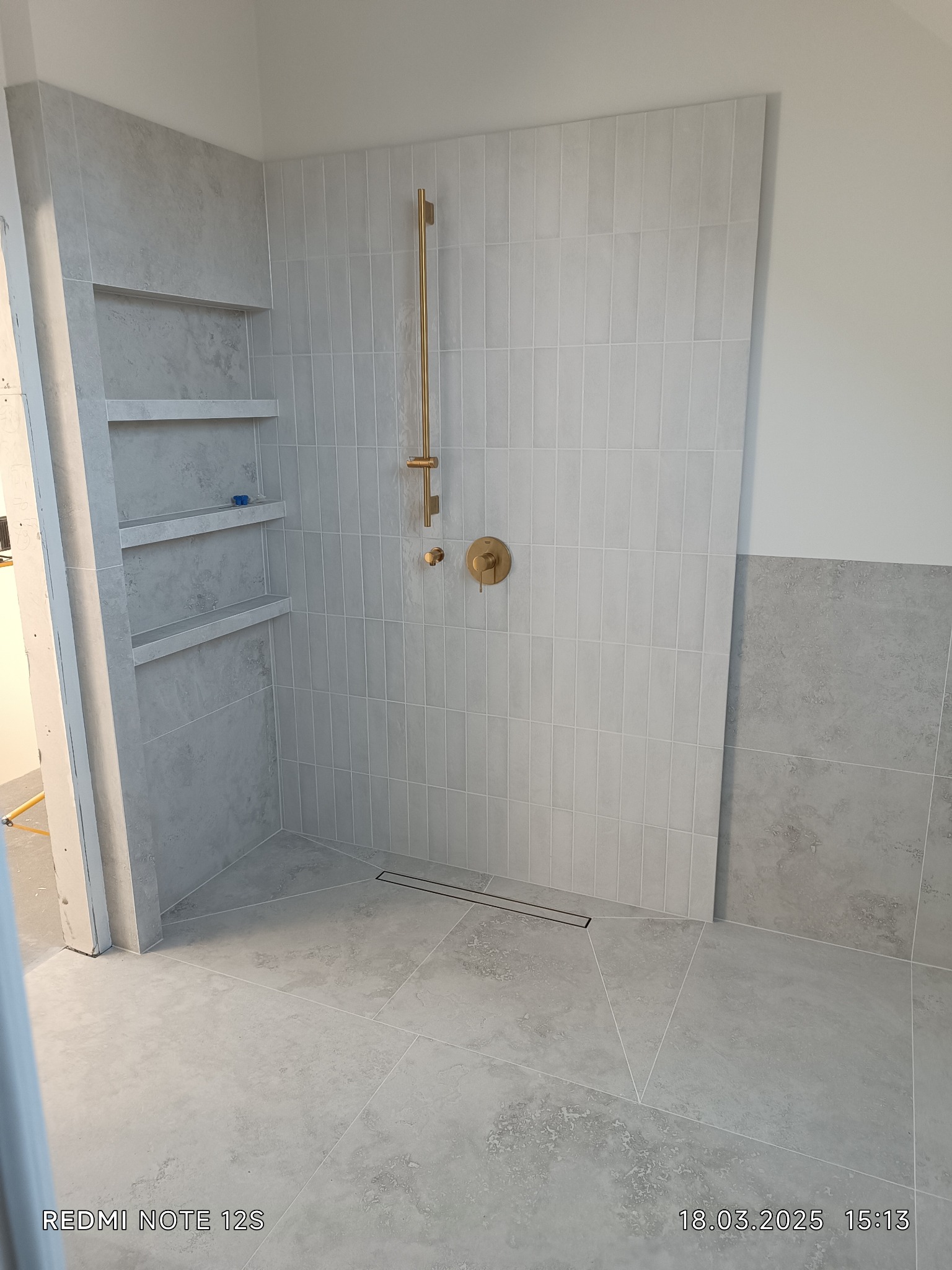 Wykończona kabina prysznicowa z szarymi płytkami, złotą armaturą i wnękami półkowymi. Widoczny odpływ liniowy i fragment ściany z dolną częścią wyłożoną płytkami.
