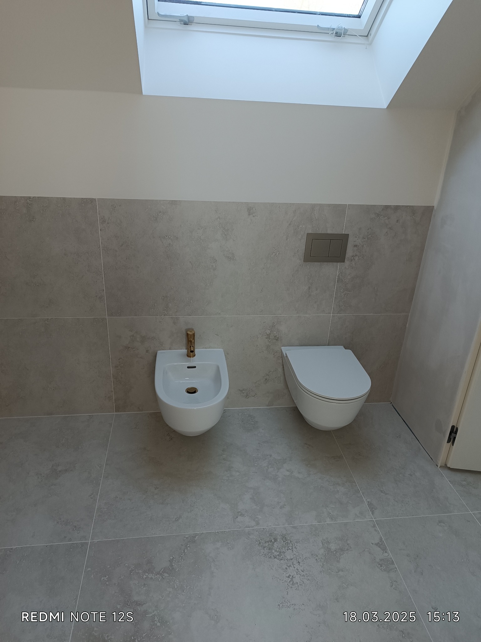 Wykończona łazienka w szarościach z podwieszaną toaletą i bidetem ze złotą armaturą. Nad sanitariatami okno dachowe wpuszczające naturalne światło.