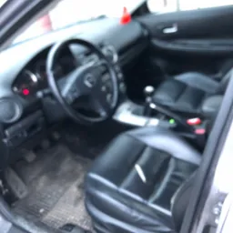 Wnętrze samochodu Mazda 6 z widocznymi czarnymi skórzanymi fotelami, deską rozdzielczą i dywanikami, wymagające prania tapicerki.