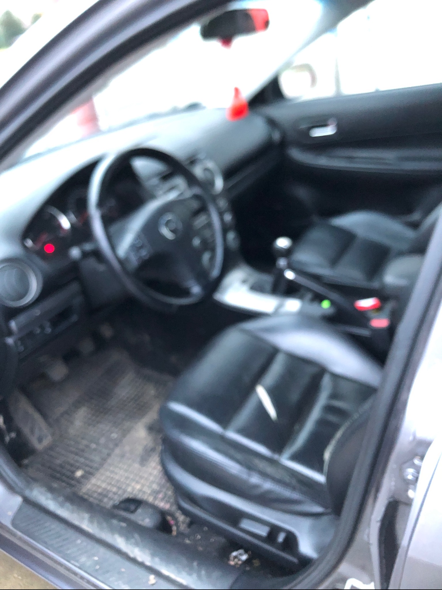 Wnętrze samochodu Mazda 6 z widocznymi czarnymi skórzanymi fotelami, deską rozdzielczą i dywanikami, wymagające prania tapicerki.