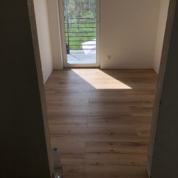 BUDIPEX OSKAR ZREDA - Pusty pokój z drewnianą podłogą w trakcie wykańczania, widok przez surowy otwór drzwiowy, butelka wody przy ścianie, światło słoneczne wpadające przez okno balkonowe.