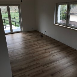 BUDIPEX OSKAR ZREDA - Pokój po remoncie z drewnopodobną podłogą, białymi ścianami, oknem z widokiem na zieleń i balkonem, z wiszącą żarówką bez klosza.