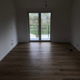 BUDIPEX OSKAR ZREDA - Pusty pokój w Szczecinie po położeniu paneli podłogowych, z widokiem na balkon i niezabezpieczoną żarówką zwisającą z sufitu.