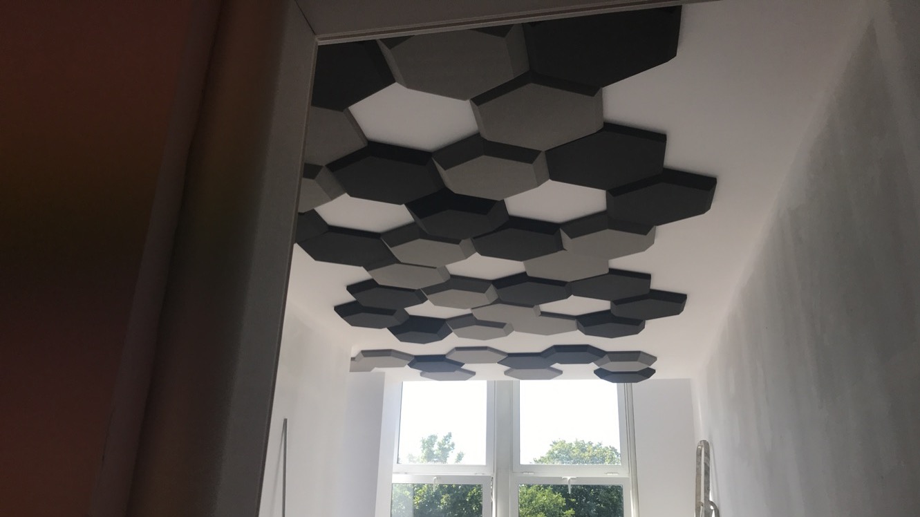 Dekoracyjny sufit z heksagonalnych paneli akustycznych w odcieniach szarości, widok z dołu na jasne okno z zielenią na zewnątrz, korytarz z częściowo widoczną ścianą