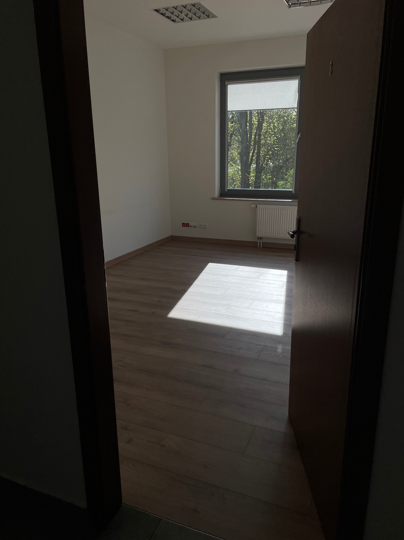 Pusty pokój z drewnianą podłogą i grzejnikiem pod oknem, widziany przez otwarte, ciemne drzwi, z wyraźnym promieniem światła słonecznego padającym na podłogę.