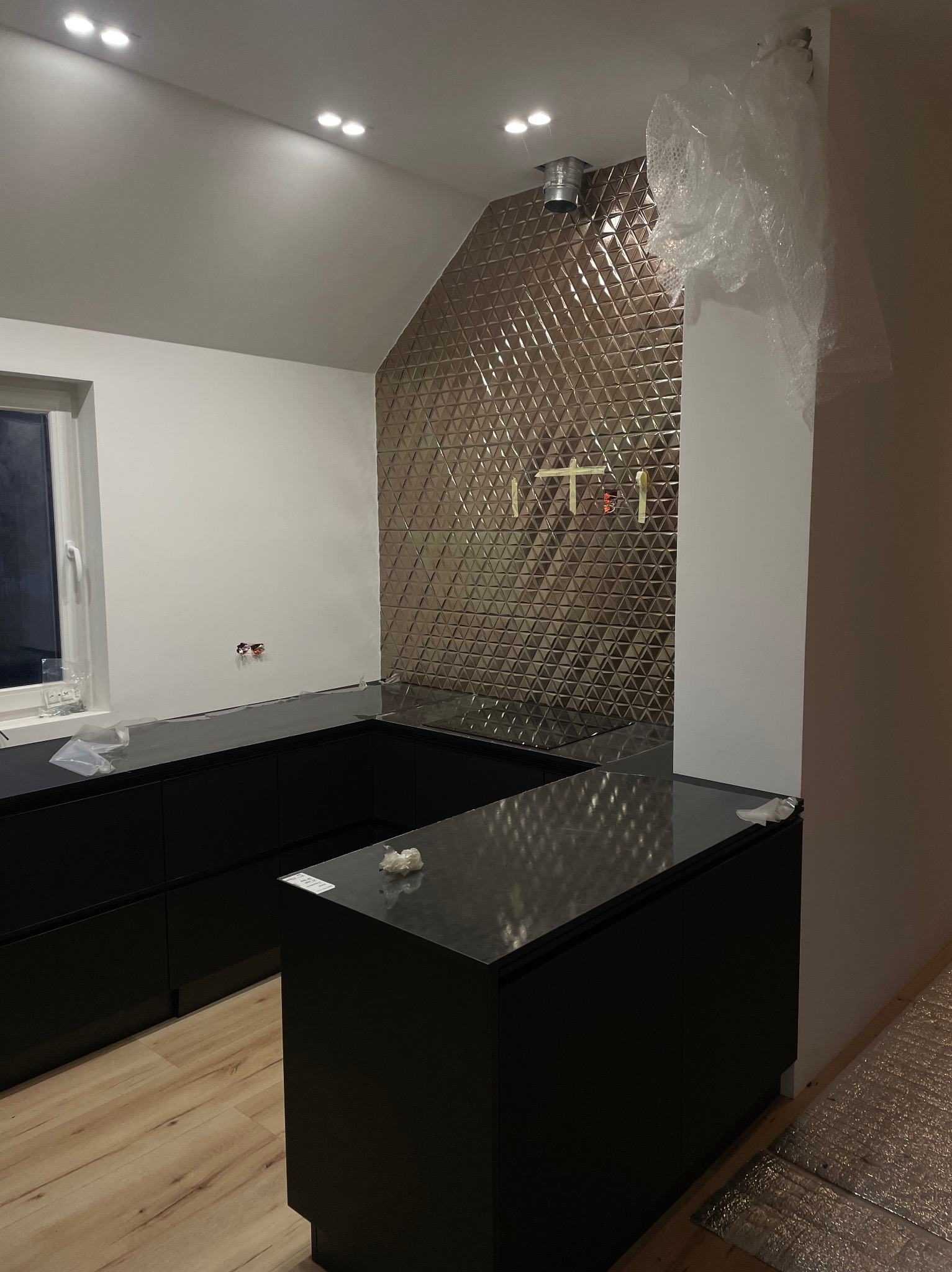 Częściowo wykończona kuchnia z czarnymi szafkami i błyszczącą, trójkątną mozaiką na ścianie, zabezpieczona folią kolumna, widoczne przygotowania do dalszych prac.