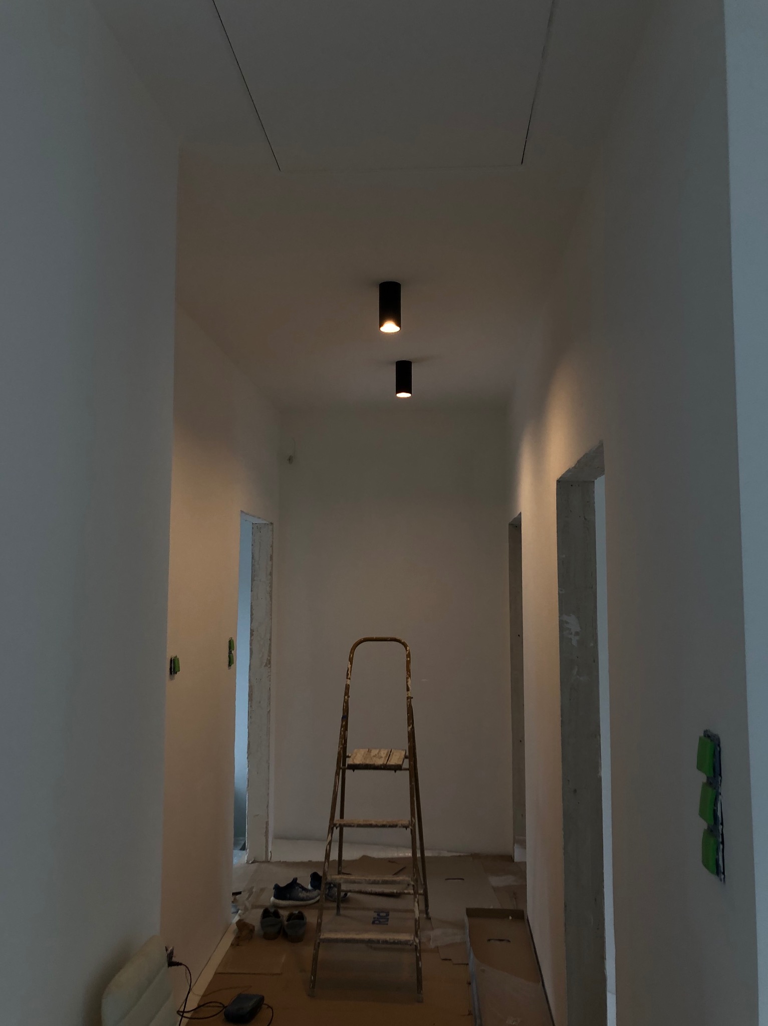 Korytarz w trakcie prac wykończeniowych: bielone ściany, drabina malarska, widoczne gniazdka elektryczne zabezpieczone taśmą, osobiste buty i sprzęt na podłodze, oraz dwa punktowe czarne reflektory...