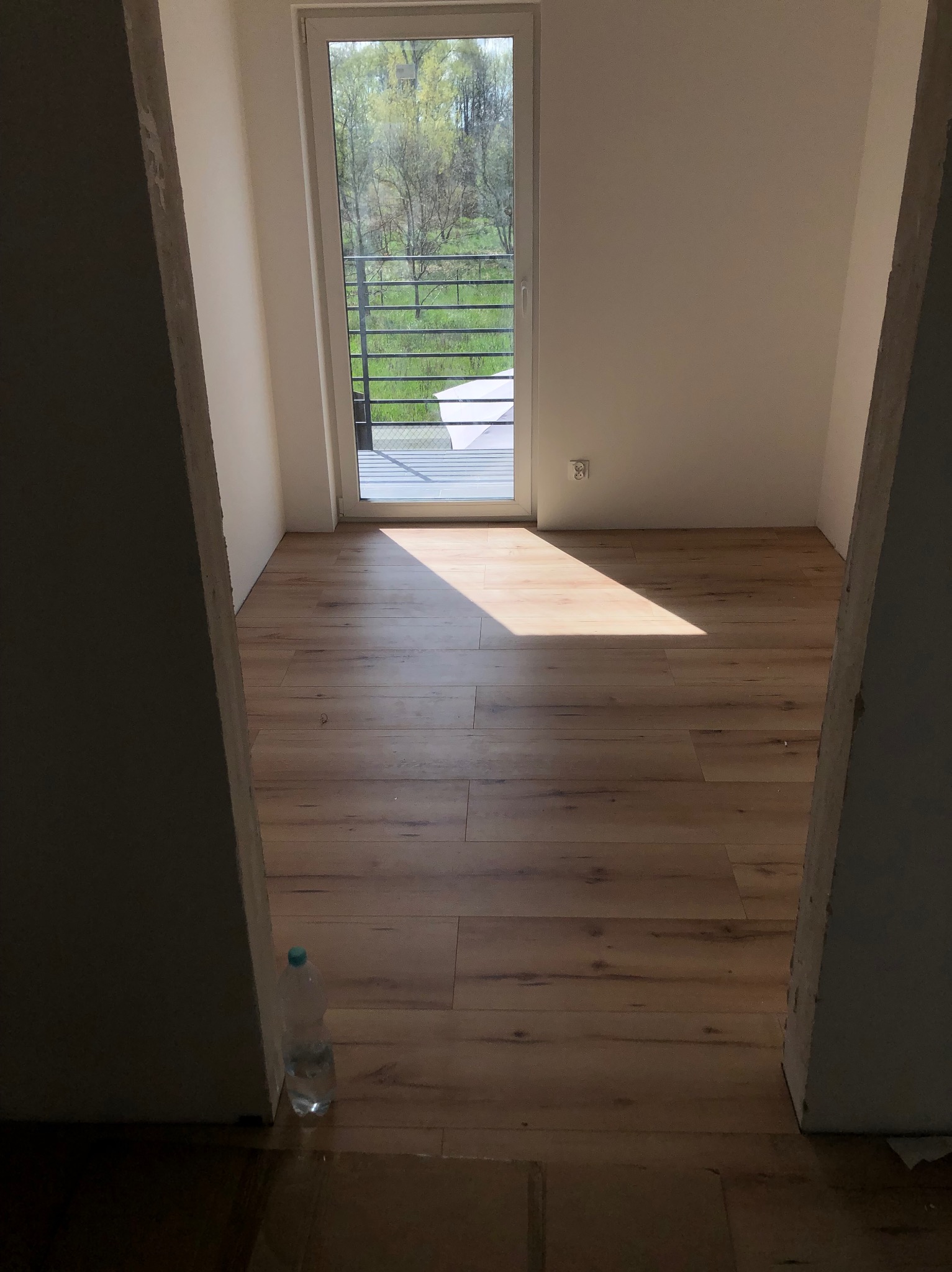 Pusty pokój z drewnianą podłogą w trakcie wykańczania, widok przez surowy otwór drzwiowy, butelka wody przy ścianie, światło słoneczne wpadające przez okno balkonowe.