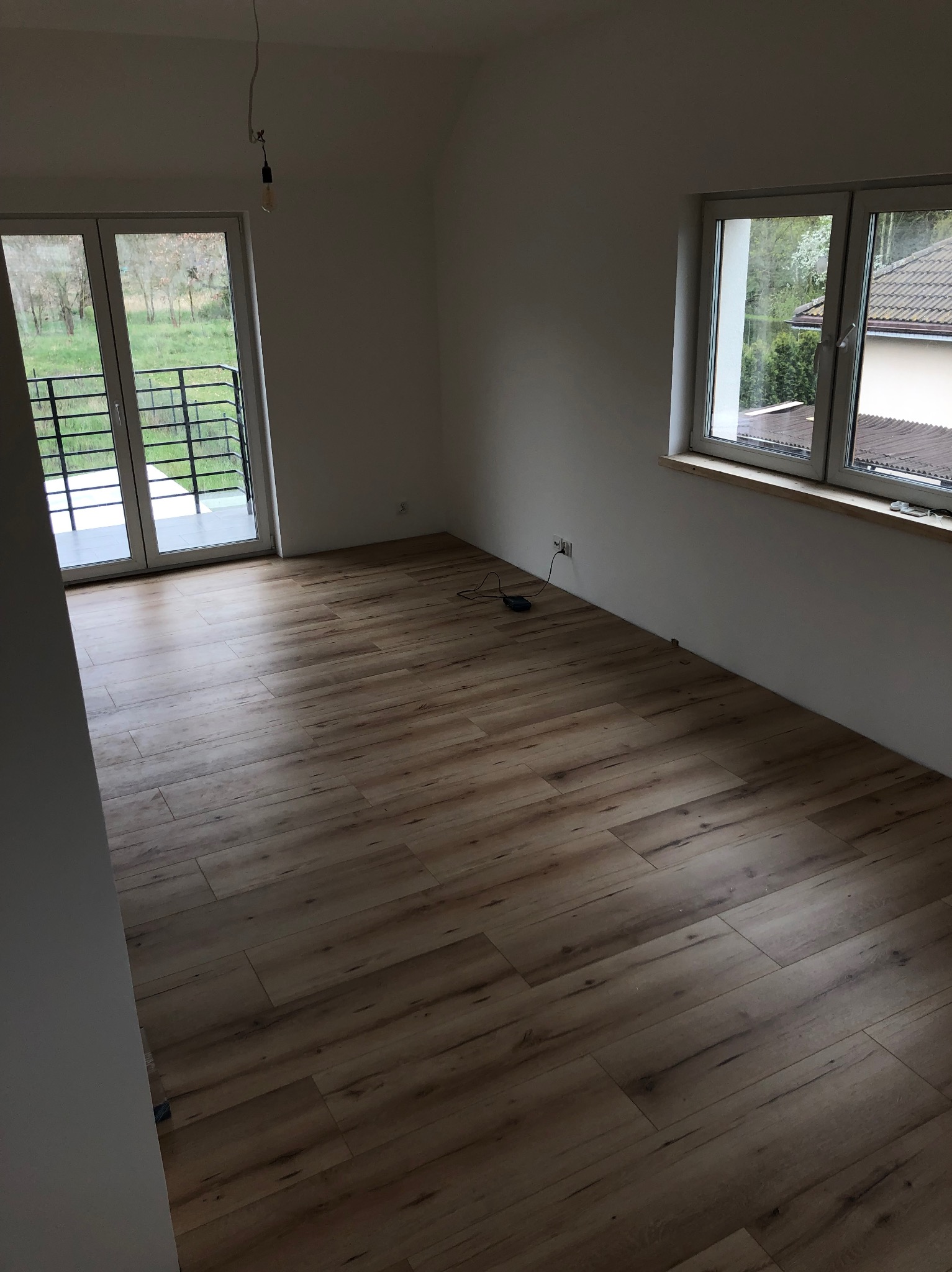 Pokój po remoncie z drewnopodobną podłogą, białymi ścianami, oknem z widokiem na zieleń i balkonem, z wiszącą żarówką bez klosza.