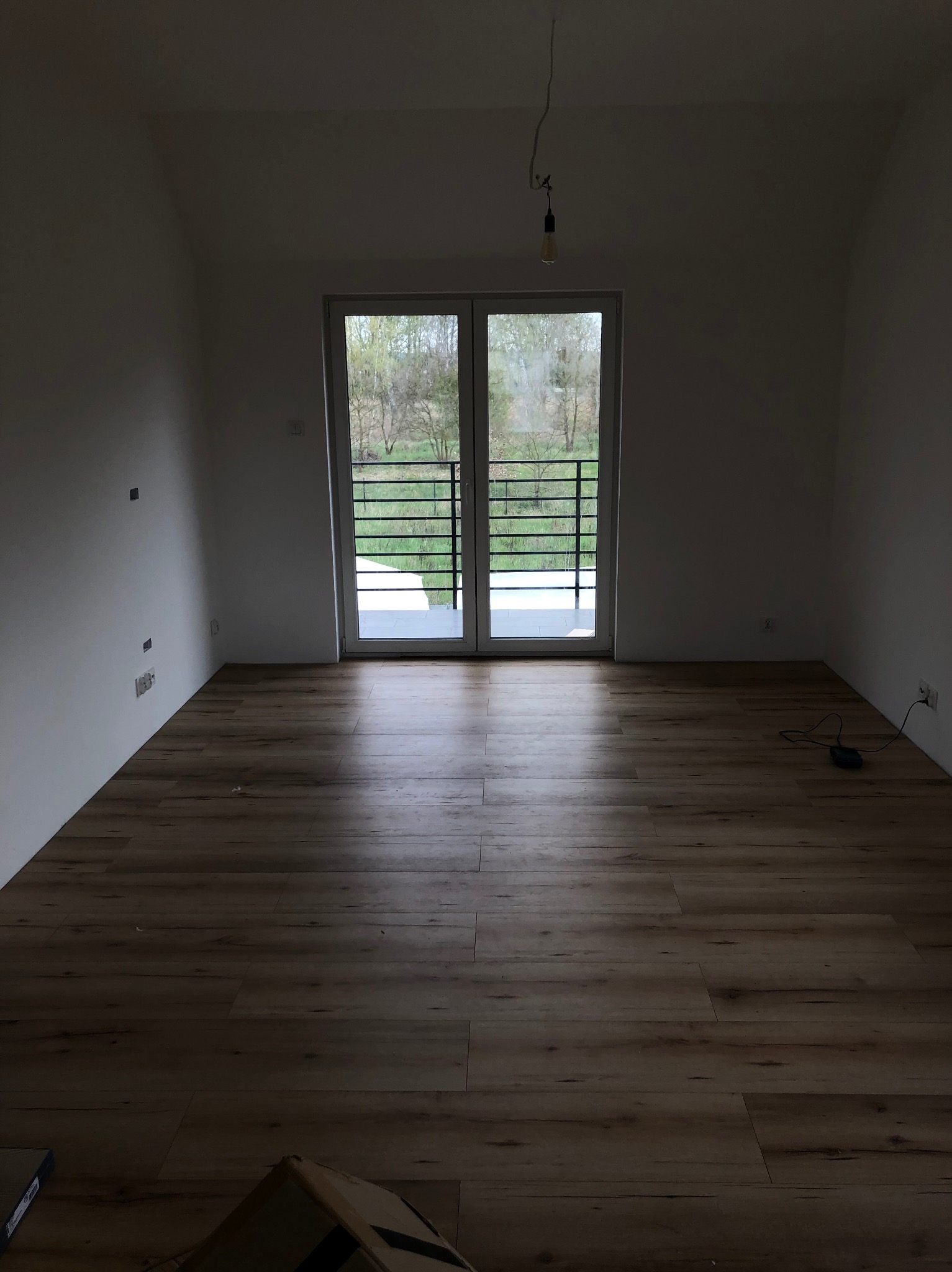 Pusty pokój w Szczecinie po położeniu paneli podłogowych, z widokiem na balkon i niezabezpieczoną żarówką zwisającą z sufitu.