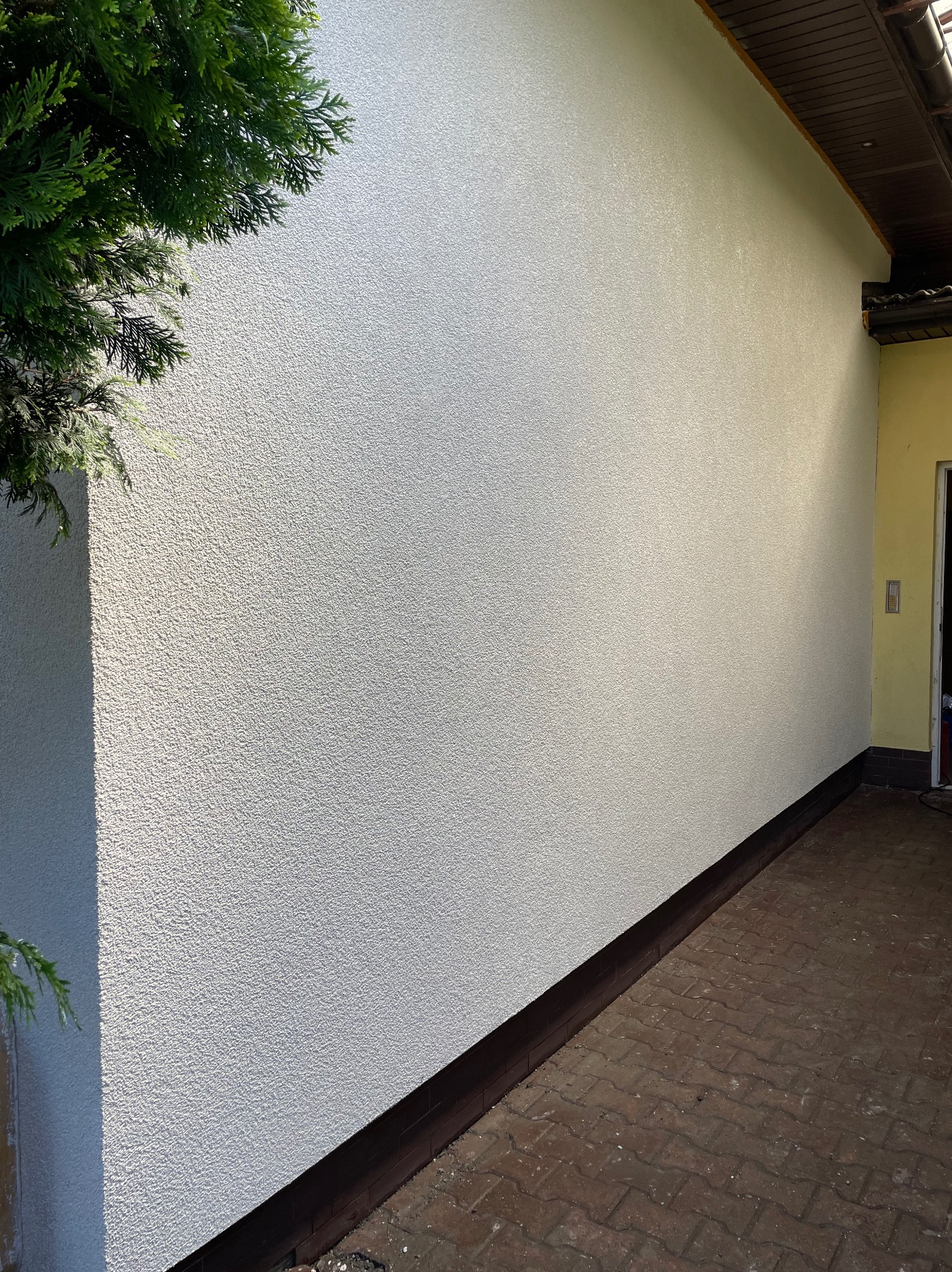 Ściana zewnętrzna z jasnym tynkiem strukturalnym, widok z boku. Częściowo widoczny fragment roślinności i brązowy cokół przy ziemi. Drobna faktura tynku.