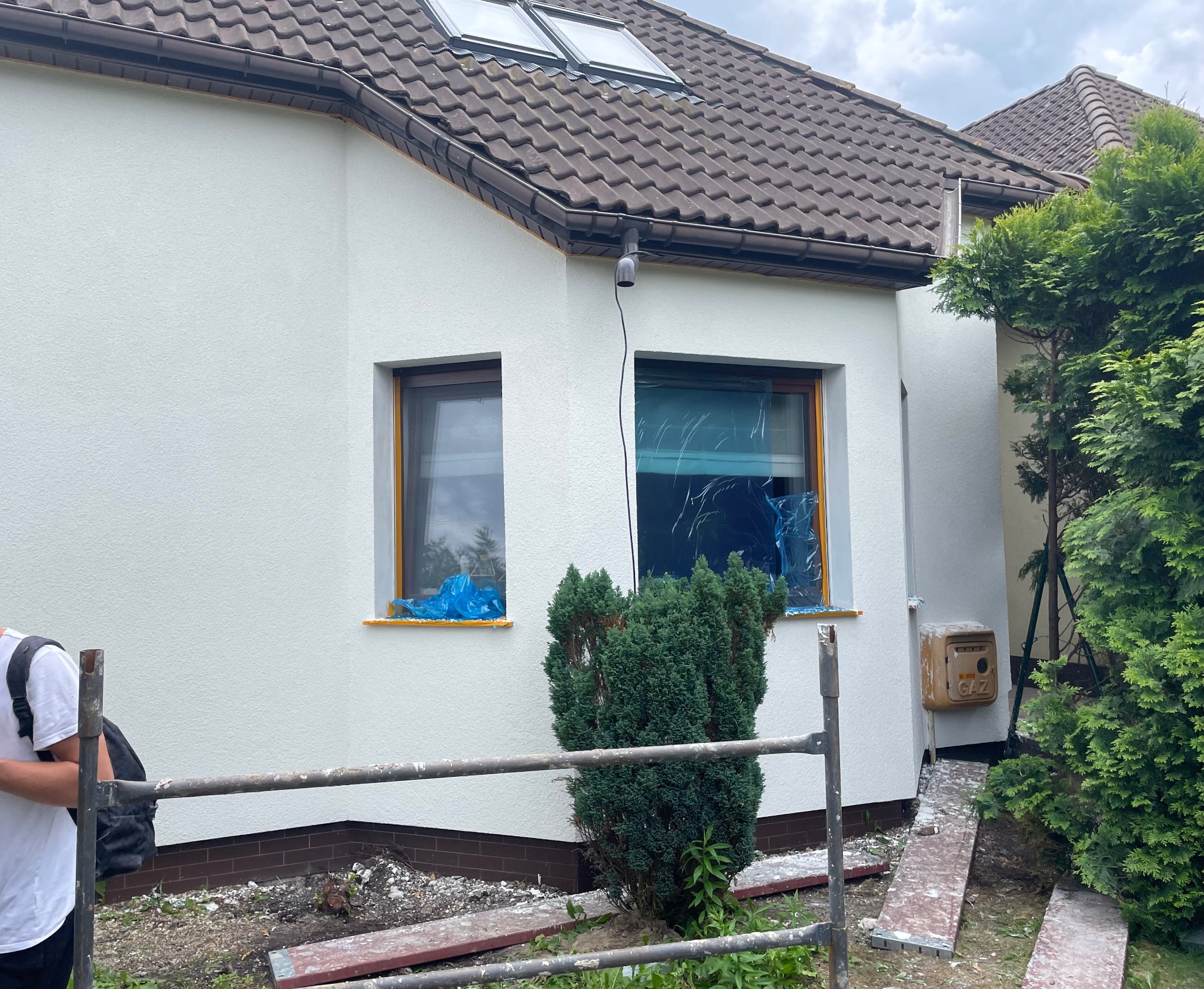 Biała elewacja domu jednorodzinnego z oknami zabezpieczonymi folią. Widoczne rusztowanie, skrzynka gazowa i fragment osoby. Prace przygotowawcze do malowania.