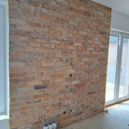 P.P. GRANIT PAWEŁ PRZĘCZEK - Ściana z cegły dekoracyjnej w trakcie montażu, zabezpieczona taśmą malarską przy oknach i drzwiach balkonowych, widoczne otwory na gniazdka elektryczne, butelki z preparatami i ściereczka...