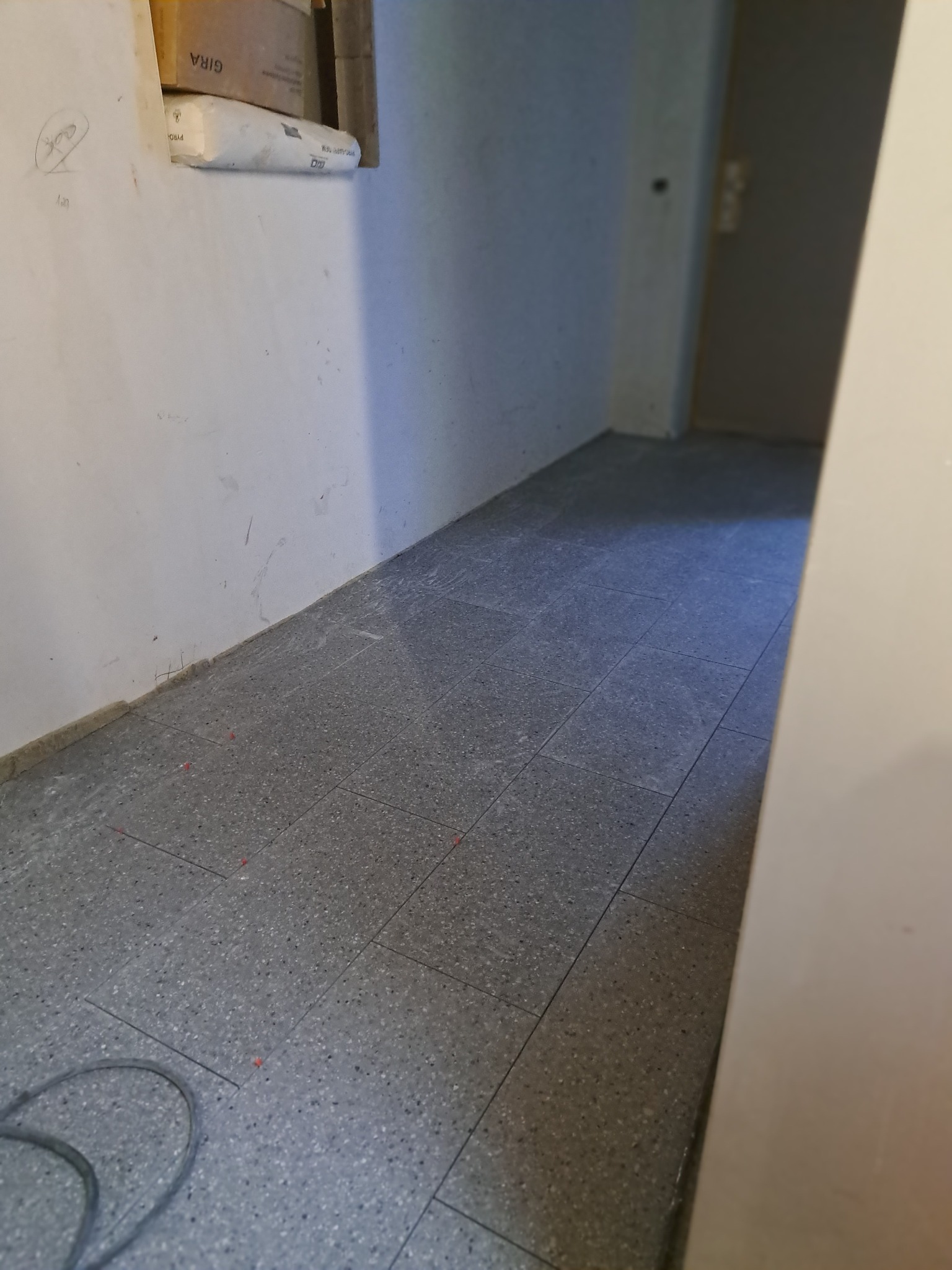 Szare płytki podłogowe ułożone w korytarzu, widoczne ślady po pracy i materiały budowlane w tle.