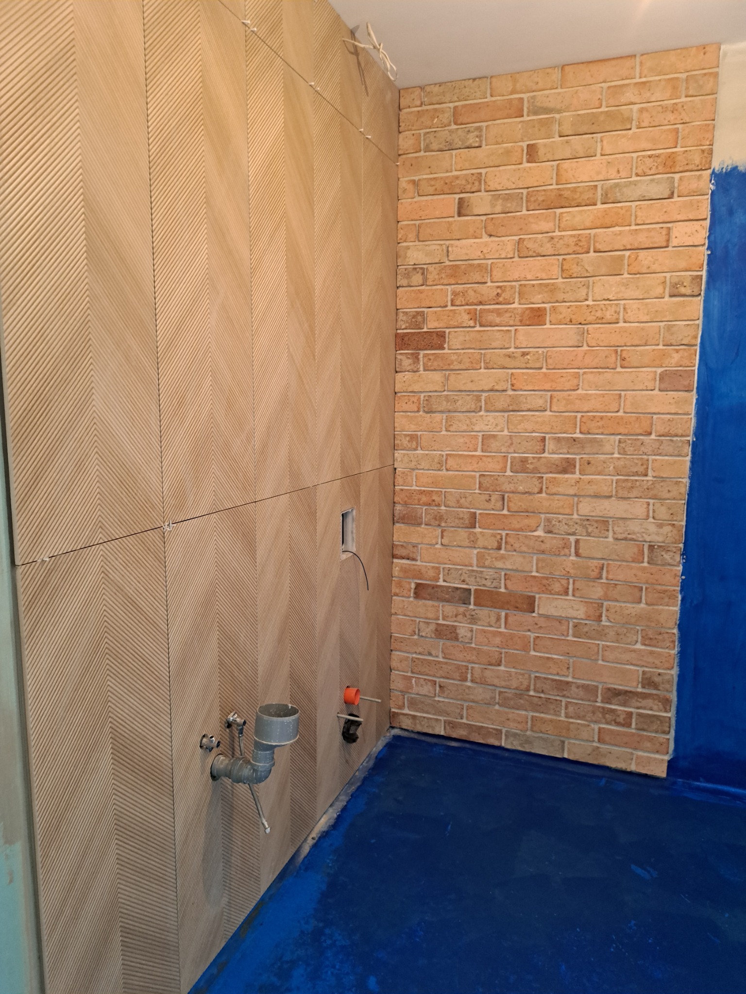 Łazienka w trakcie prac wykończeniowych: ściana z płytek ułożonych we wzór jodełki, fragment ściany wyłożony cegłą, zabezpieczona folią podłoga i wystające rury odpływowe.