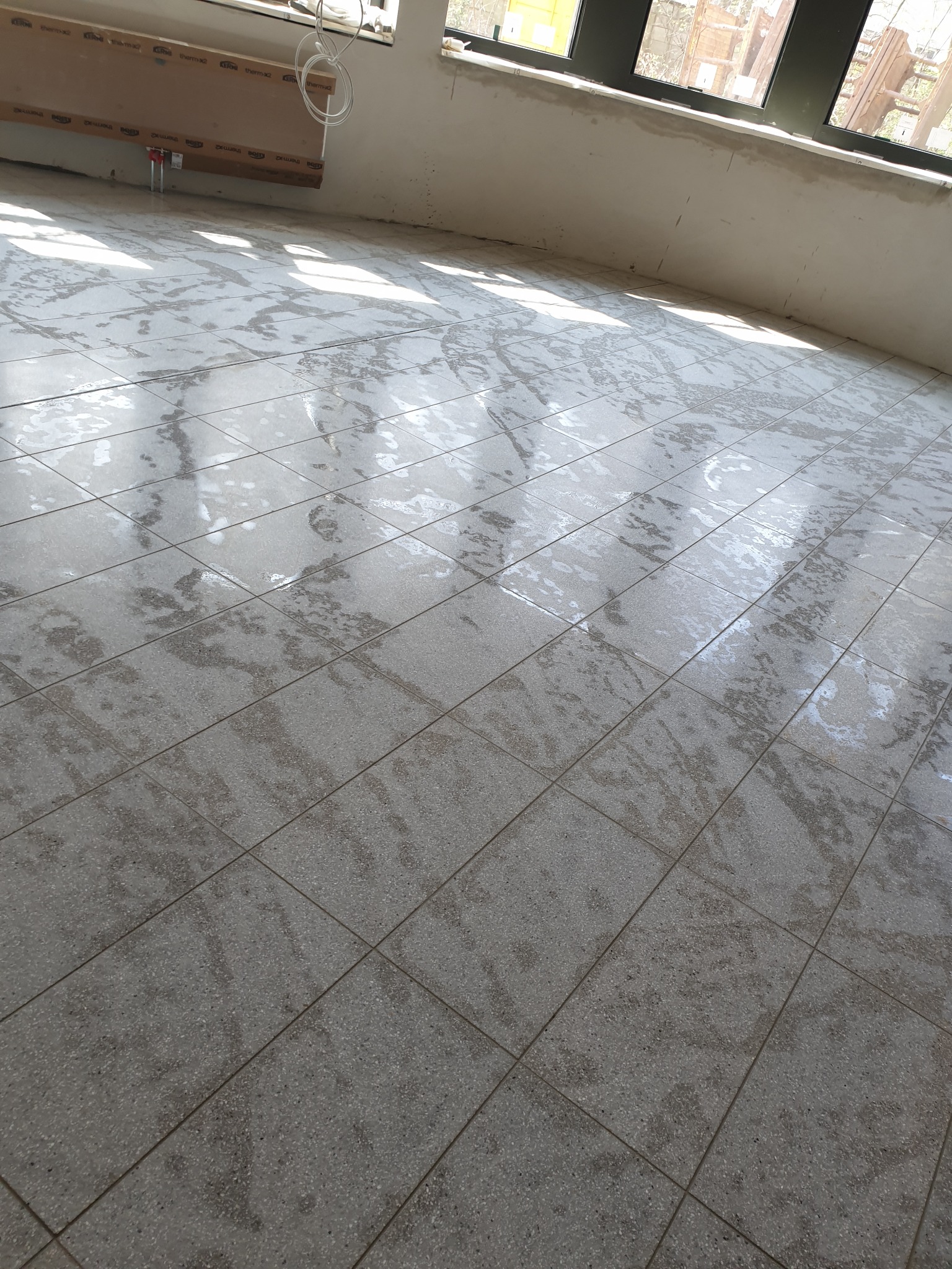 Świeżo ułożona, szara, terakotowa podłoga w pomieszczeniu w trakcie remontu, z widocznymi promieniami słonecznymi wpadającymi przez okno i instalacją grzewczą na ścianie.