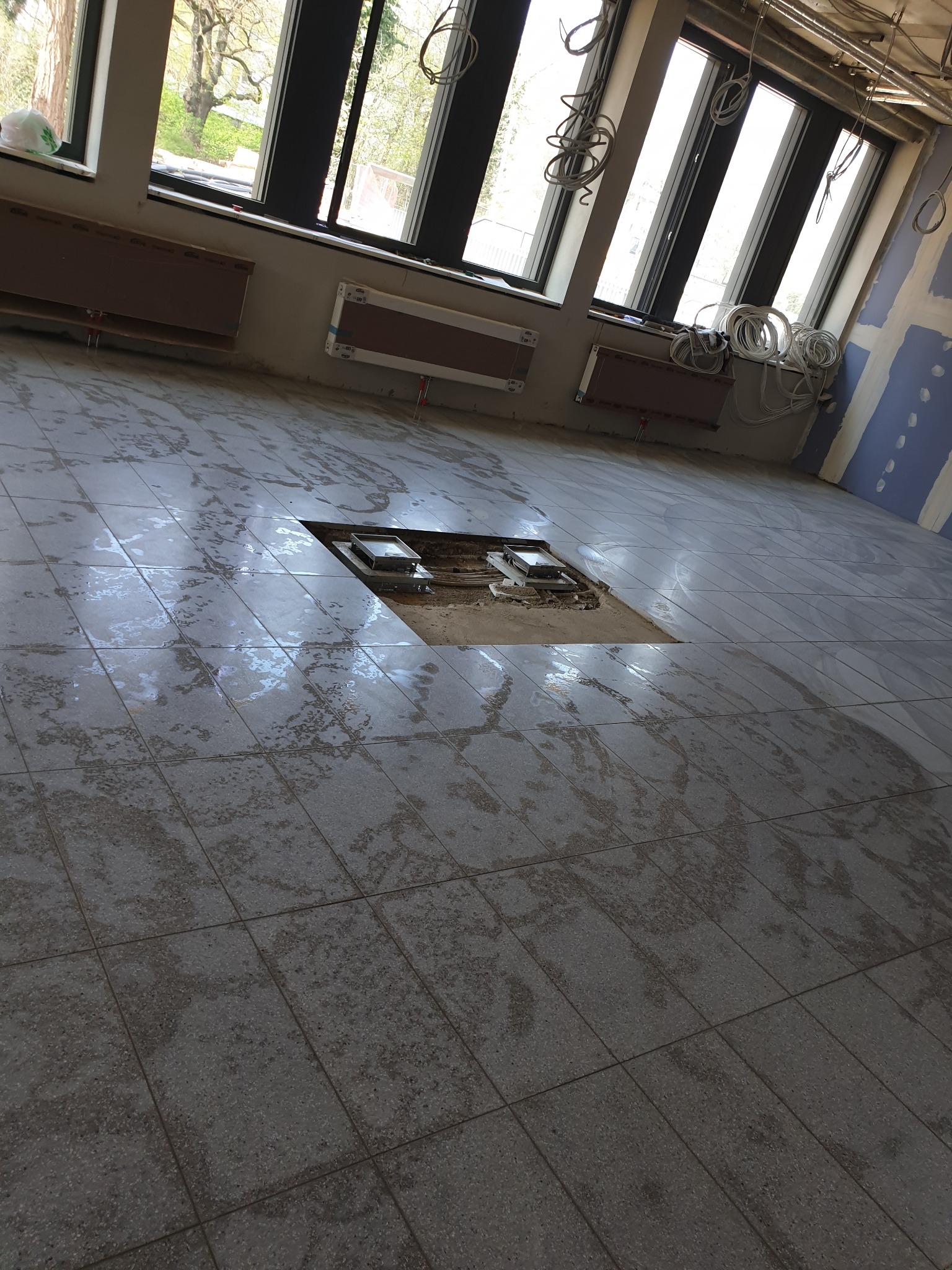 Wnętrze w trakcie remontu z położoną szarą terakotą, widoczne kable zwisające z sufitu, ślady wilgoci na podłodze i otwór w posadzce z instalacją.