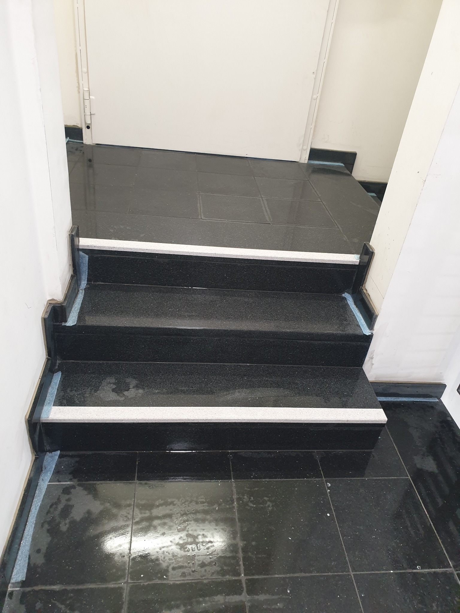 Granitowe schody wewnętrzne z białymi paskami antypoślizgowymi, wykończone czarną fugą, widoczne zabezpieczenia folią malarską.