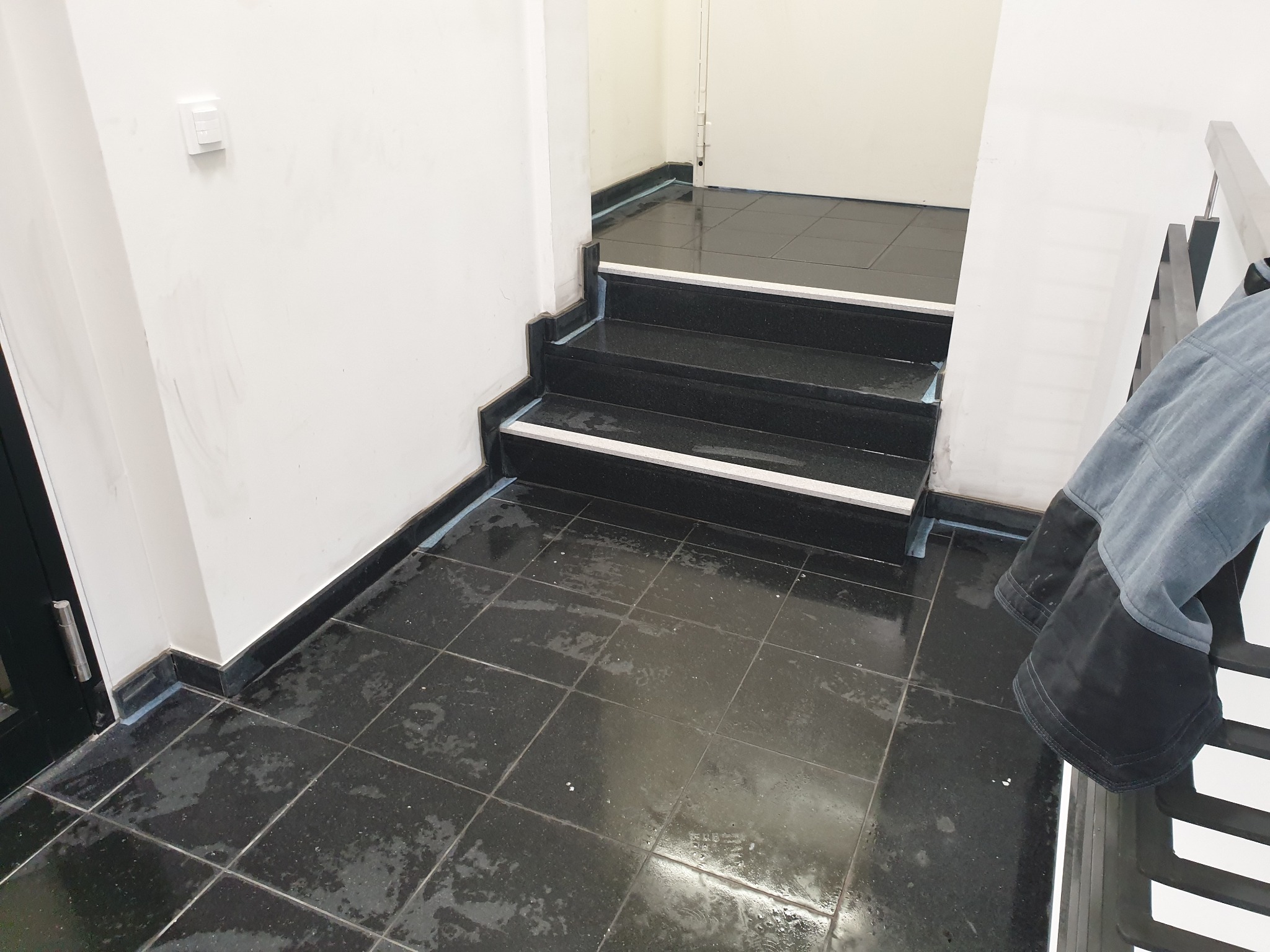 Błyszczące, czarne płytki podłogowe i schody z białymi akcentami, prawdopodobnie po świeżym umyciu, z widoczną krawędzią schodów i fragmentem ubrania wiszącego na poręczy.