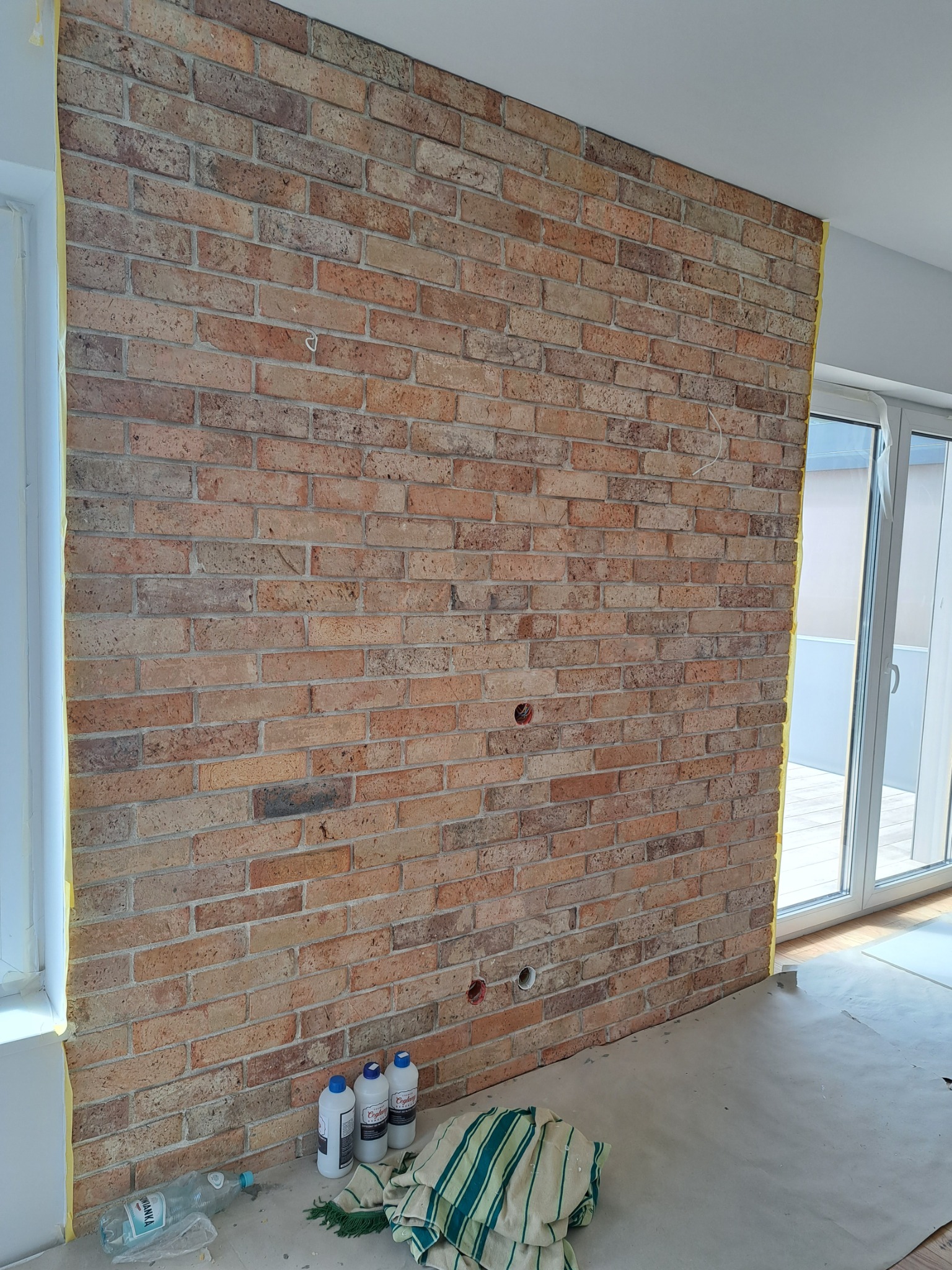 Ściana z cegły dekoracyjnej w trakcie montażu, zabezpieczona taśmą malarską przy oknach i drzwiach balkonowych, widoczne otwory na gniazdka elektryczne, butelki z preparatami i ściereczka...