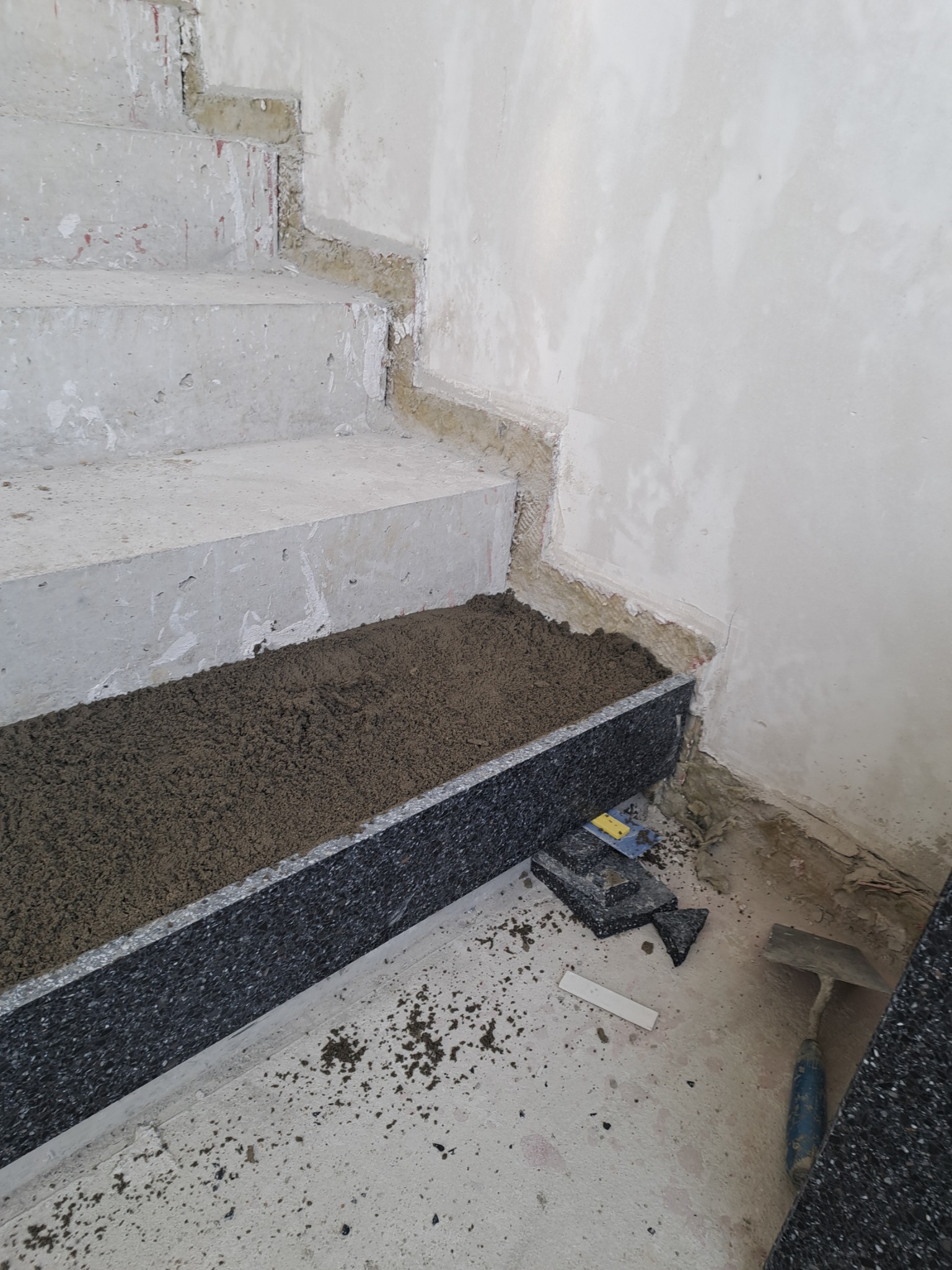 Przygotowanie do montażu okładziny schodów: wypełniona zaprawą krawędź stopnia z widoczną kielnią i fragmentami okładziny na podłodze w surowym stanie.