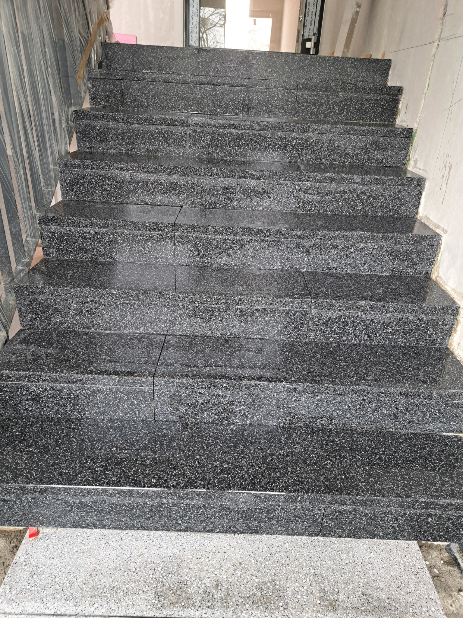 Schody wewnętrzne wyłożone ciemnym kamieniem kompozytowym o drobnej fakturze, widoczne połączenia między płytami, dolny stopień w jasnym odcieniu, zabezpieczone folią ściany w tle.