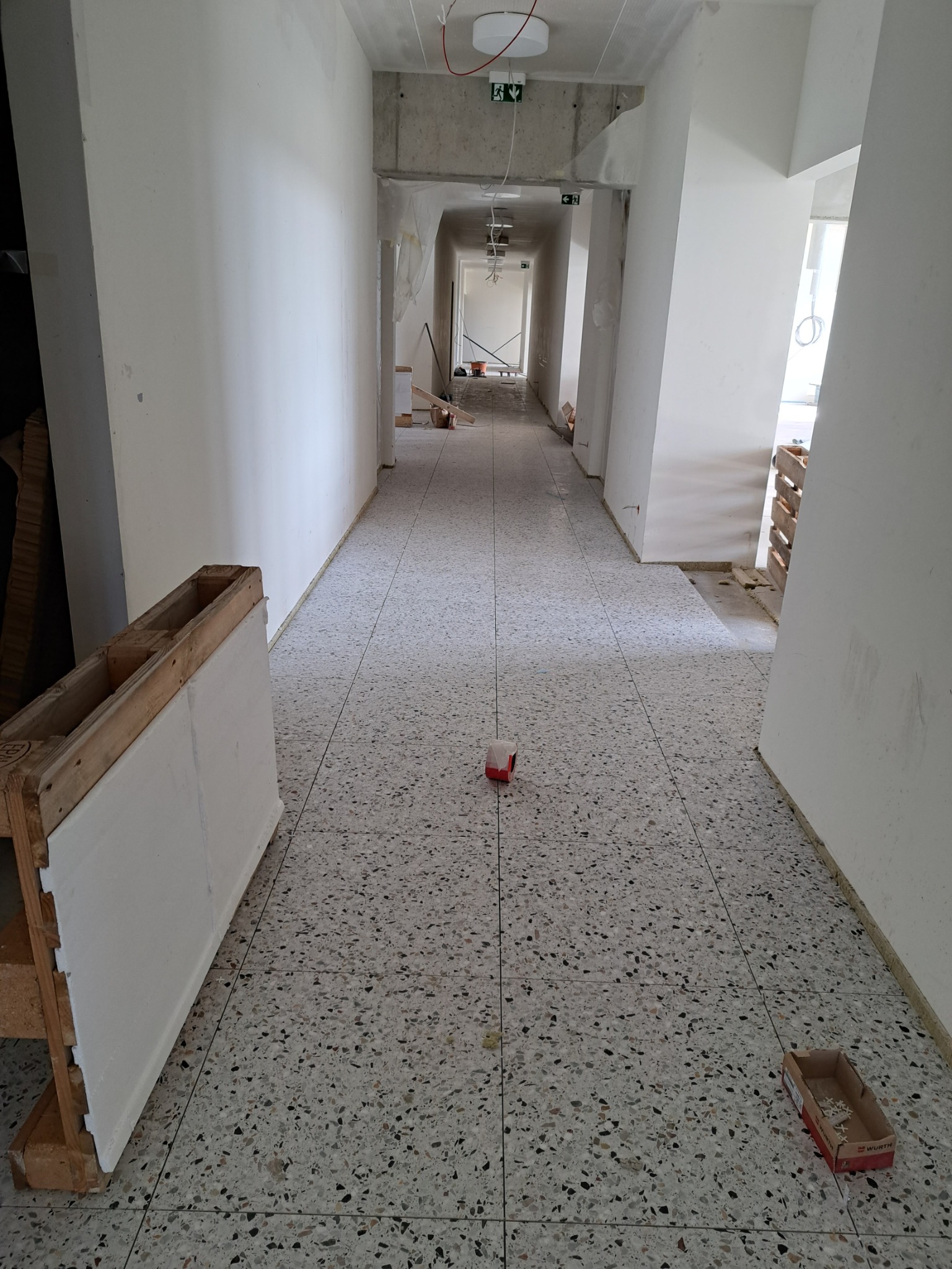 Korytarz w trakcie prac wykończeniowych, świeżo pomalowane ściany na biało, widoczne elementy konstrukcyjne sufitu, podłoga wyłożona płytkami lastryko, paleta z płytą kartonowo-gipsową oraz pudełko...