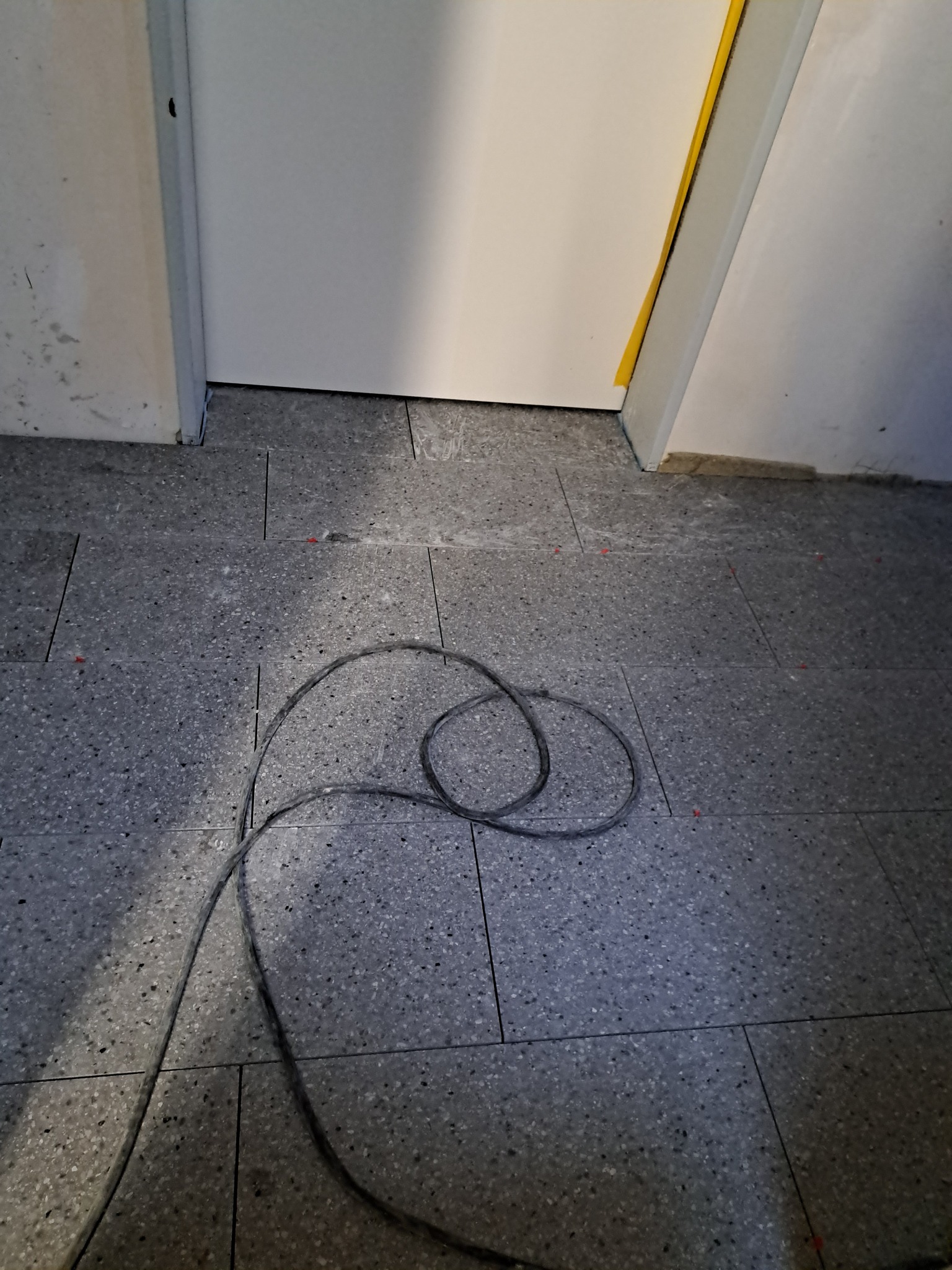 Szara, kamienna podłoga z widocznymi spoinami i fragmentem kabla. Czerwone kliny dystansowe pomiędzy płytkami. Widoczny próg drzwi z żółtą taśmą zabezpieczającą.
