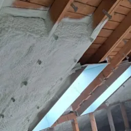 Poddasze ocieplone pianką PUR, widoczne krokwie i metalowe uchwyty mocujące, jasna piana pokrywająca skośną powierzchnię.