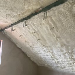 PROFI-PUR IHOR VOLKOV - Poddasze po natrysku izolacji piankowej, widoczne metalowe elementy mocujące. Biała, nierówna powierzchnia piany pokrywa skośny sufit i fragment ściany.