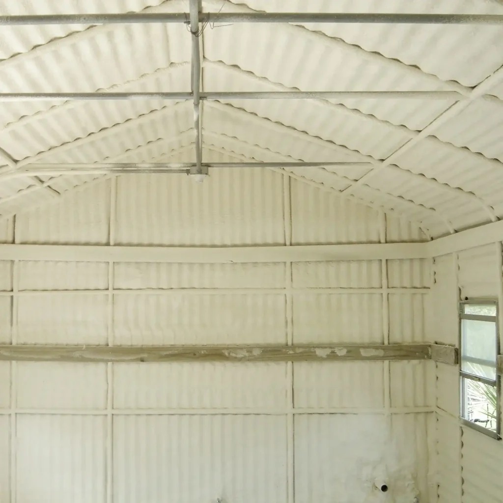 Wnętrze budynku z metalową konstrukcją dachu i ścian, pokryte warstwą jasnej pianki izolacyjnej, widoczne okno w prawej ścianie.