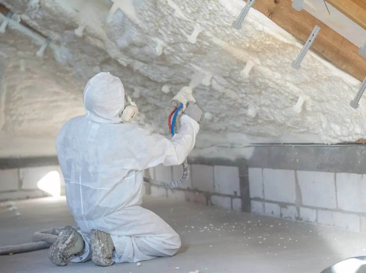 Pracownik w kombinezonie ochronnym aplikuje piankę PUR na poddasze, widoczne krople pianki w powietrzu i drewniane elementy konstrukcji dachu.