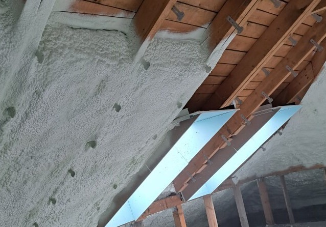 Poddasze ocieplone pianką PUR, widoczne krokwie i metalowe uchwyty mocujące, jasna piana pokrywająca skośną powierzchnię.