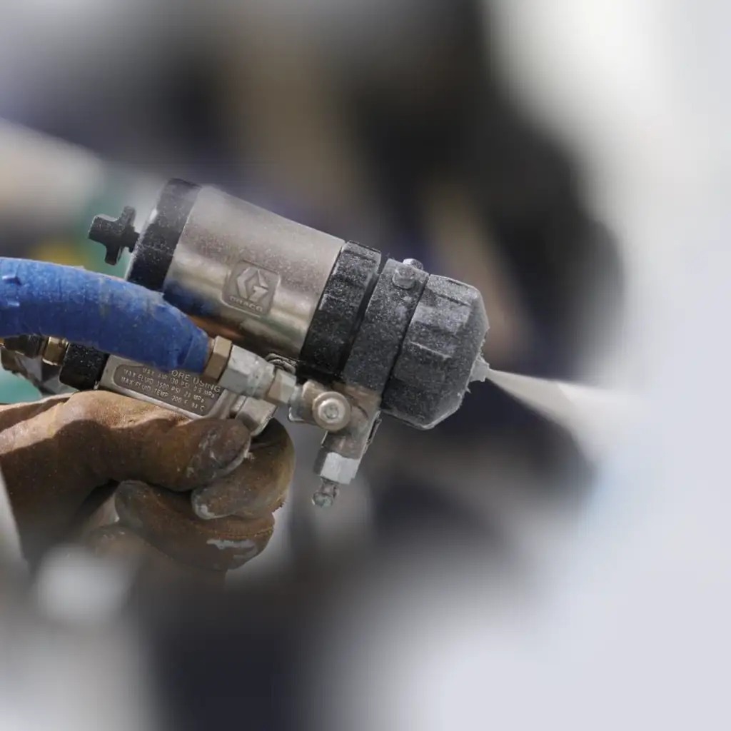 Pistolet natryskowy marki Graco w dłoni w roboczej rękawicy aplikuje pianę izolacyjną na jasną powierzchnię.