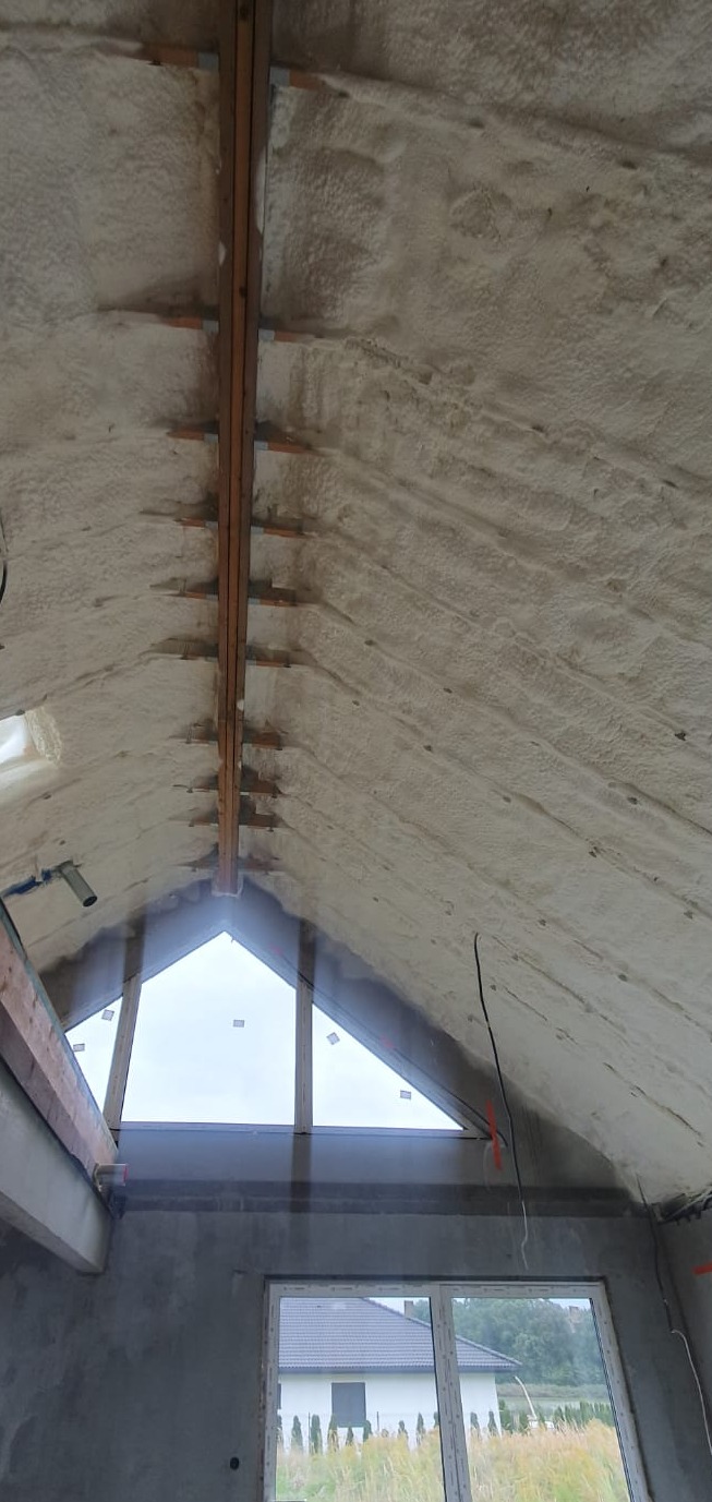 Ocieplony poddasze pianą, widoczne krokwie i okno dachowe. Biała, równomierna warstwa izolacji termicznej na skośnym dachu. Detal wykończenia poddasza.