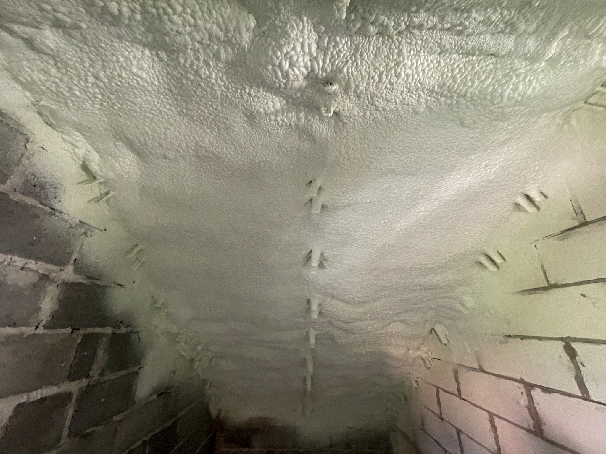 Ocieplenie pianką PUR poddasza z widocznymi elementami mocującymi, na tle ściany z cegieł. Aplikacja pianki o jednolitej strukturze, widok z dołu.