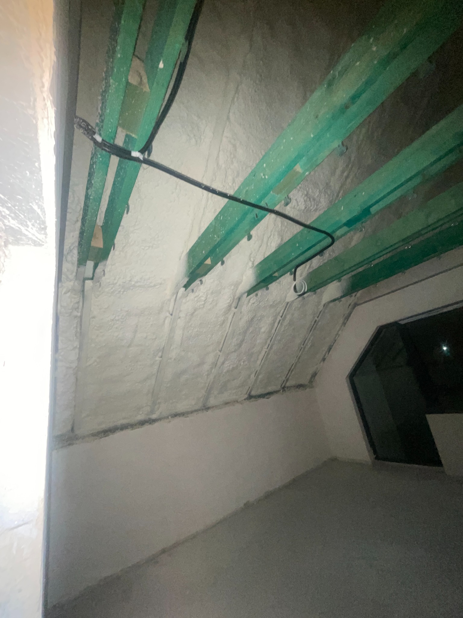 Poddasze po natrysku izolacji, widoczne zielone krokwie i instalacja elektryczna. Biała, równomierna warstwa izolacji pokrywa skośne powierzchnie dachu.