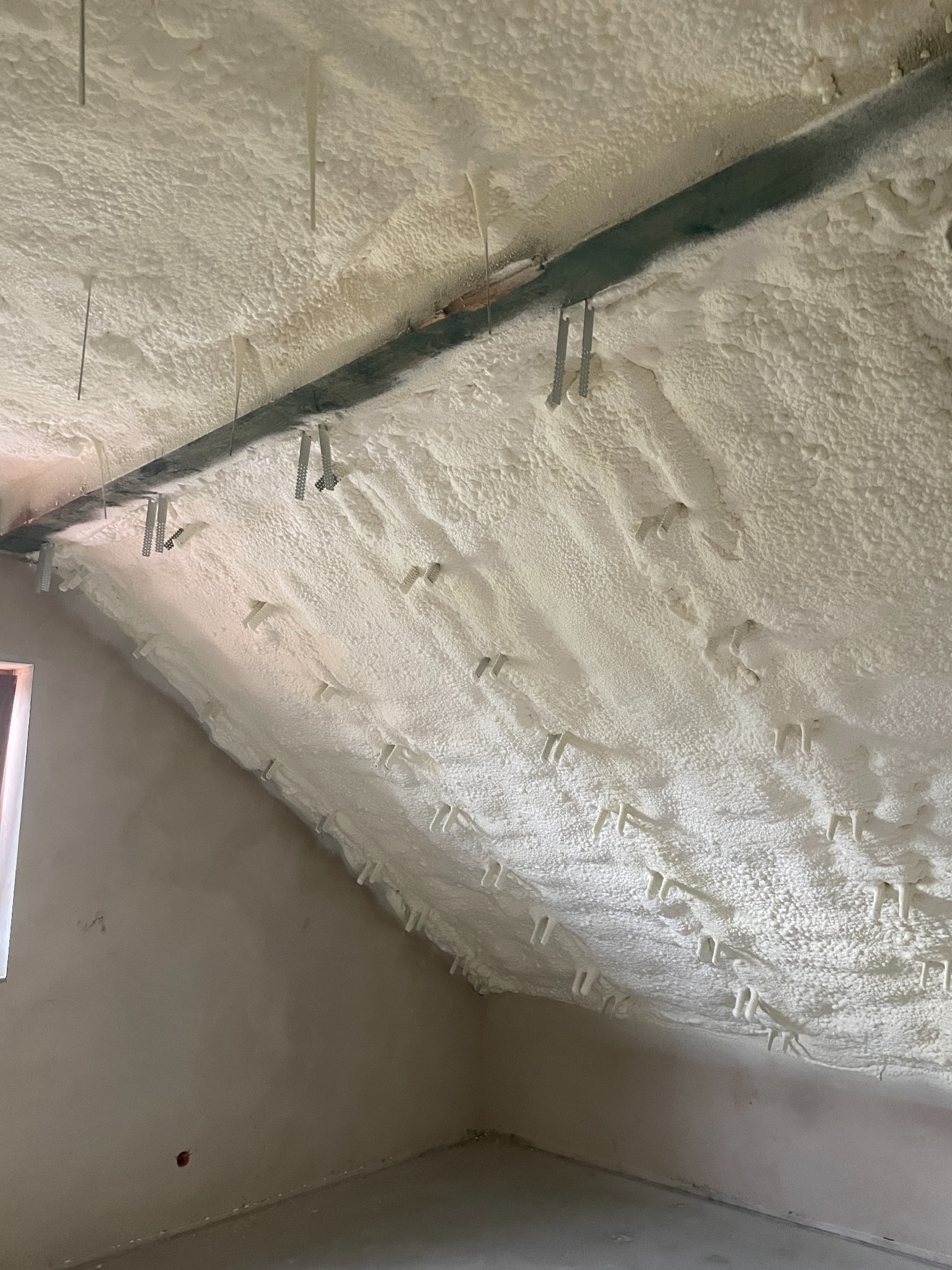 Poddasze po natrysku izolacji piankowej, widoczne metalowe elementy mocujące. Biała, nierówna powierzchnia piany pokrywa skośny sufit i fragment ściany.