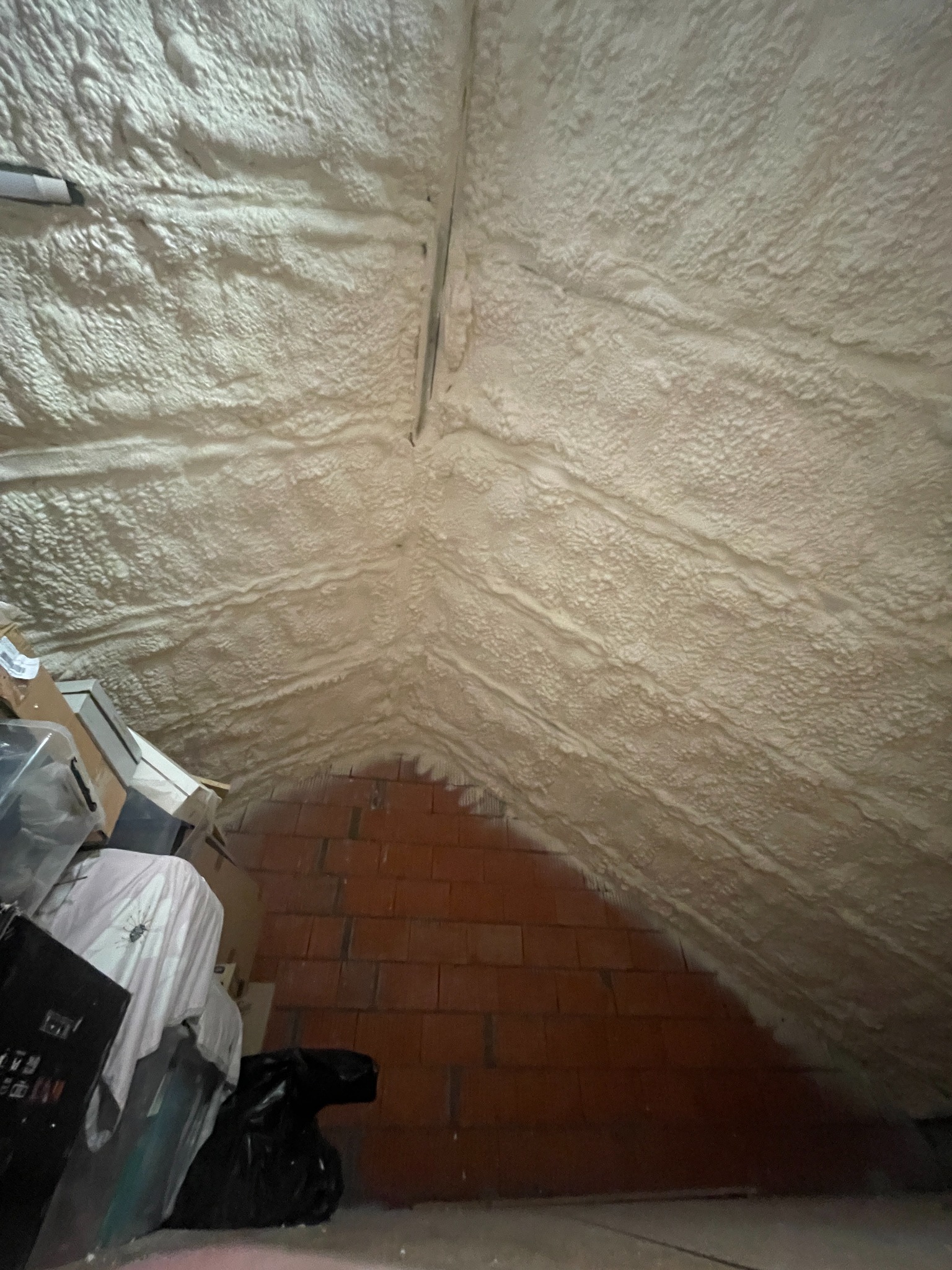 Poddasze po natrysku izolacji piankowej, widoczne cegły i kartony. Izolacja pokrywa skośny dach, tworząc jednolitą warstwę. Detal wykończenia izolacji.