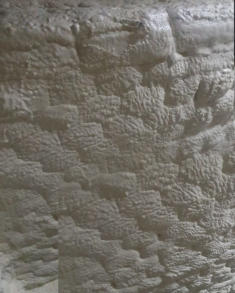 Ściana pokryta grubą warstwą białej pianki PUR, tworzącej nieregularną, pofałdowaną strukturę. Widoczne nierówności i charakterystyczna tekstura izolacji natryskowej.