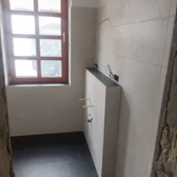 Wnętrze remontowanej łazienki z oknem, jasnymi płytkami ściennymi, ciemną podłogą i elementami instalacji wodnej wystającymi ze ściany pod umywalkę.