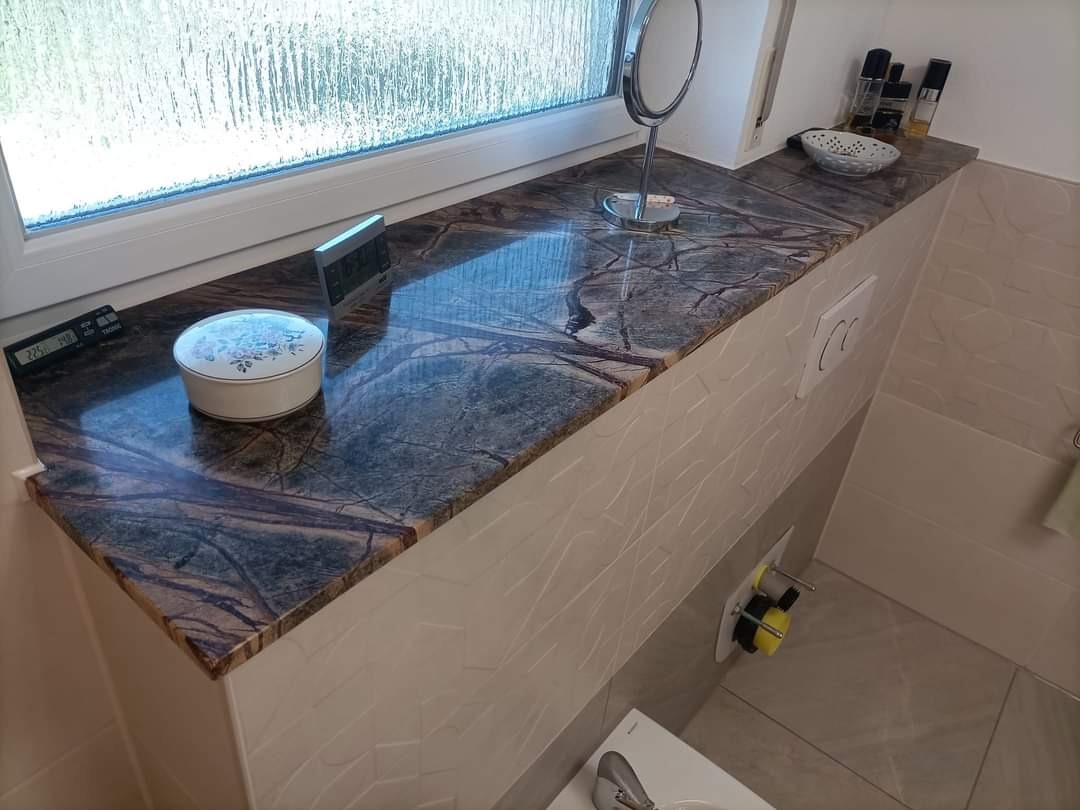 Wykończona łazienka z blatem z ciemnego marmuru pod oknem, na którym stoją dekoracje i kosmetyki, a na ścianie widoczna jest podtynkowa spłuczka toaletowa.