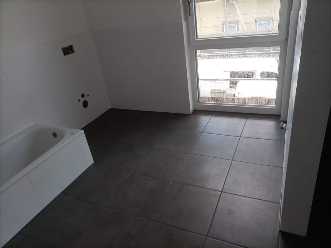 Łazienka w trakcie remontu: ułożone ciemnoszare płytki podłogowe, biała wanna, ściany częściowo wyłożone białymi płytkami, widoczne przygotowane punkty instalacji wodnej, okno z widokiem na zewnątrz.