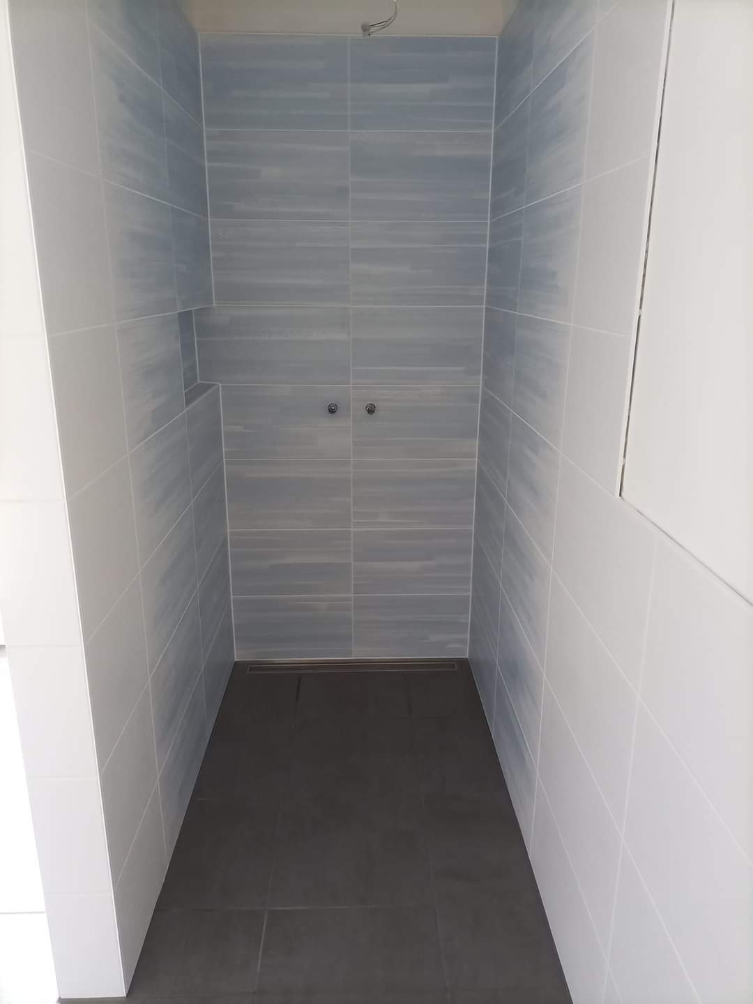 Wykończony prysznic typu walk-in z szarymi płytkami imitującymi drewno na ścianach i ciemnoszarą podłogą, widoczne dwa zawory na ścianie.