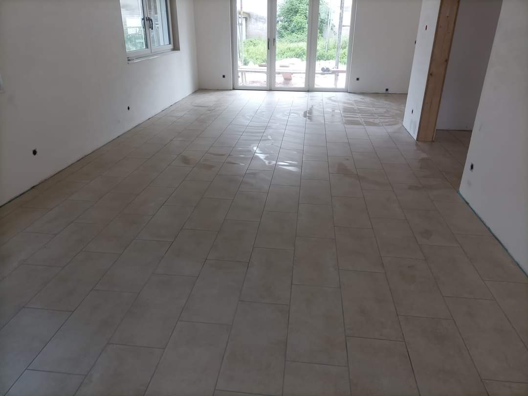 Ułożone jasne, prostokątne płytki podłogowe w nowym pomieszczeniu, widoczne ślady mokrego kleju lub czyszczenia, duże okno i drzwi balkonowe w tle.